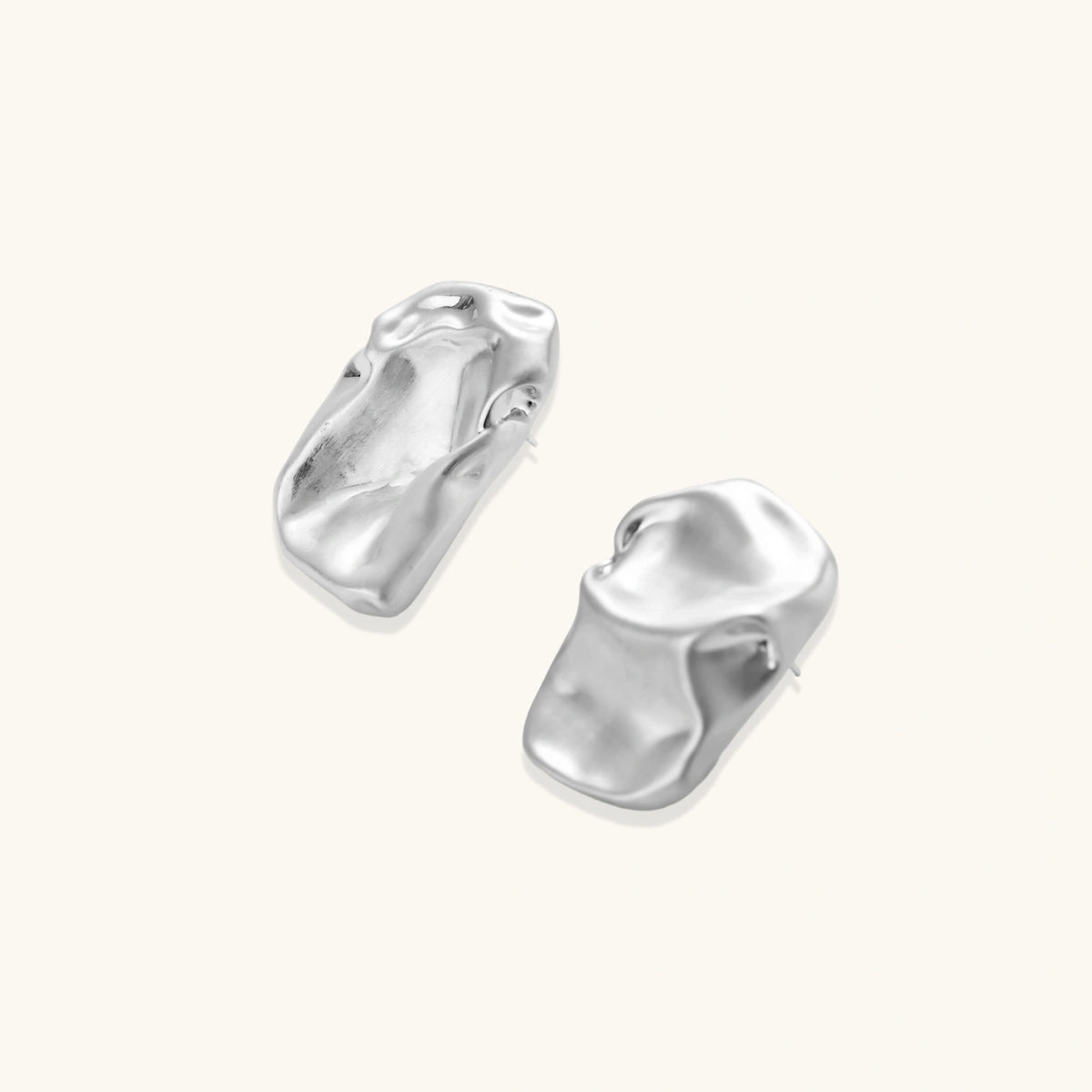 Pair of sculptural silver ear cuffs with organic fluid shapes and polished finish against cream background.